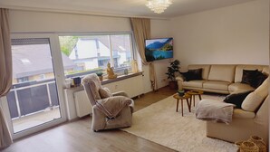 A spacious living room with a large window, a sofa, a chair, a small table, and a television. - Henno's Apartment (Bielefeld)