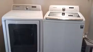 Två vita Samsung-tvättmaskiner i ett tvättrum med ett kaklat golv och en handduksställning på väggen.