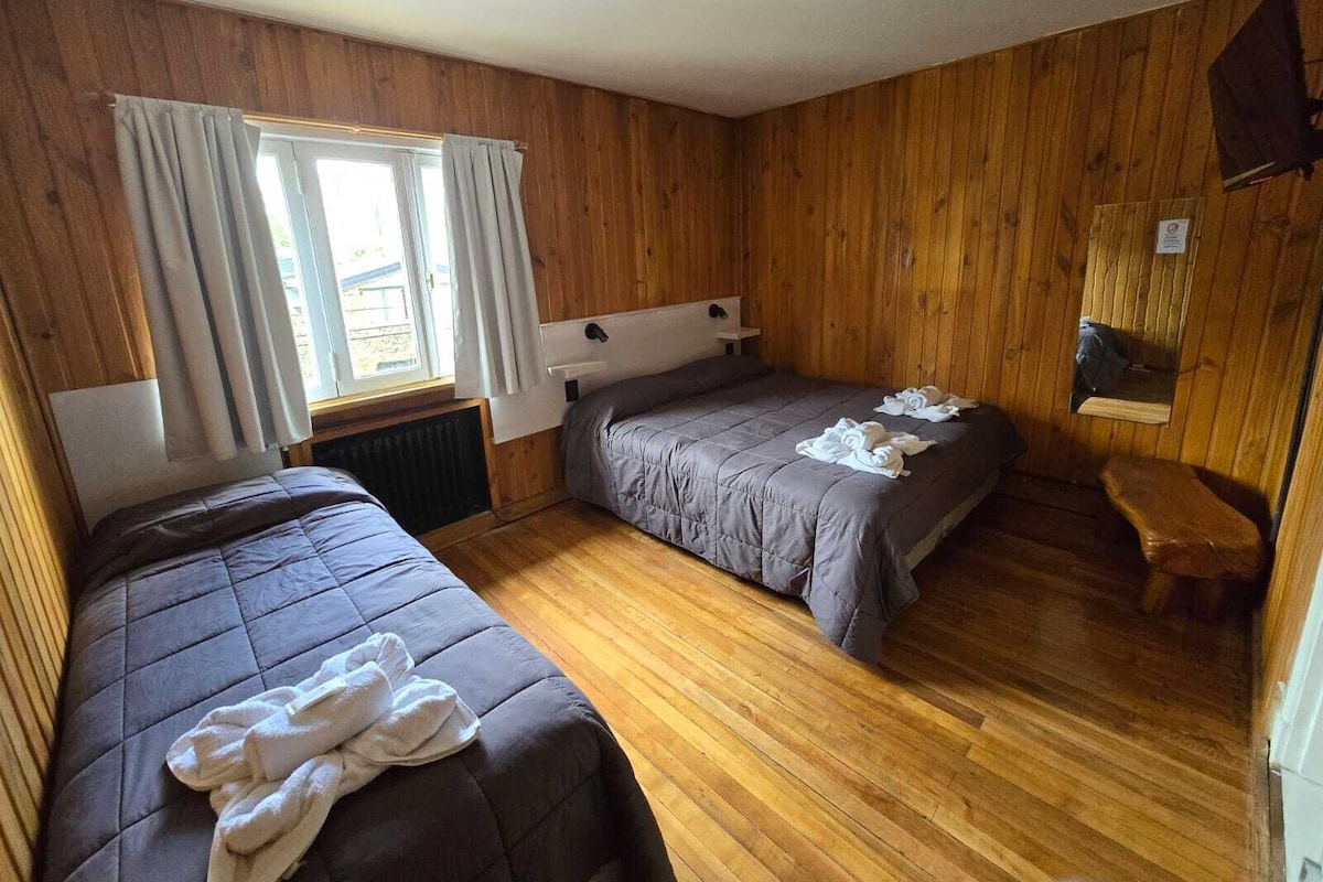 A room with two beds, wooden walls, a window with curtains, a small bench, and a mirror on the wall.
