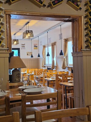 L’intérieur d’un restaurant avec des tables et des chaises en bois, des suspensions décoratives et une vue sur l’extérieur par une porte.