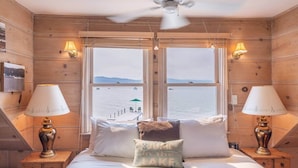 Ein Zimmer mit einem Bett, zwei Nachttischlampen, einem Deckenventilator und Seeblick durch große Fenster.