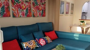 A modern living room with a teal sofa, colorful cushions, a small wooden table with a plant, and a kitchen area in the background. - Your best vacations in Mijas (Mijas)