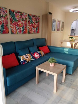 A modern living room with a teal sofa, colorful cushions, a small wooden table with a plant, and a kitchen area in the background. - Your best vacations in Mijas (Mijas)