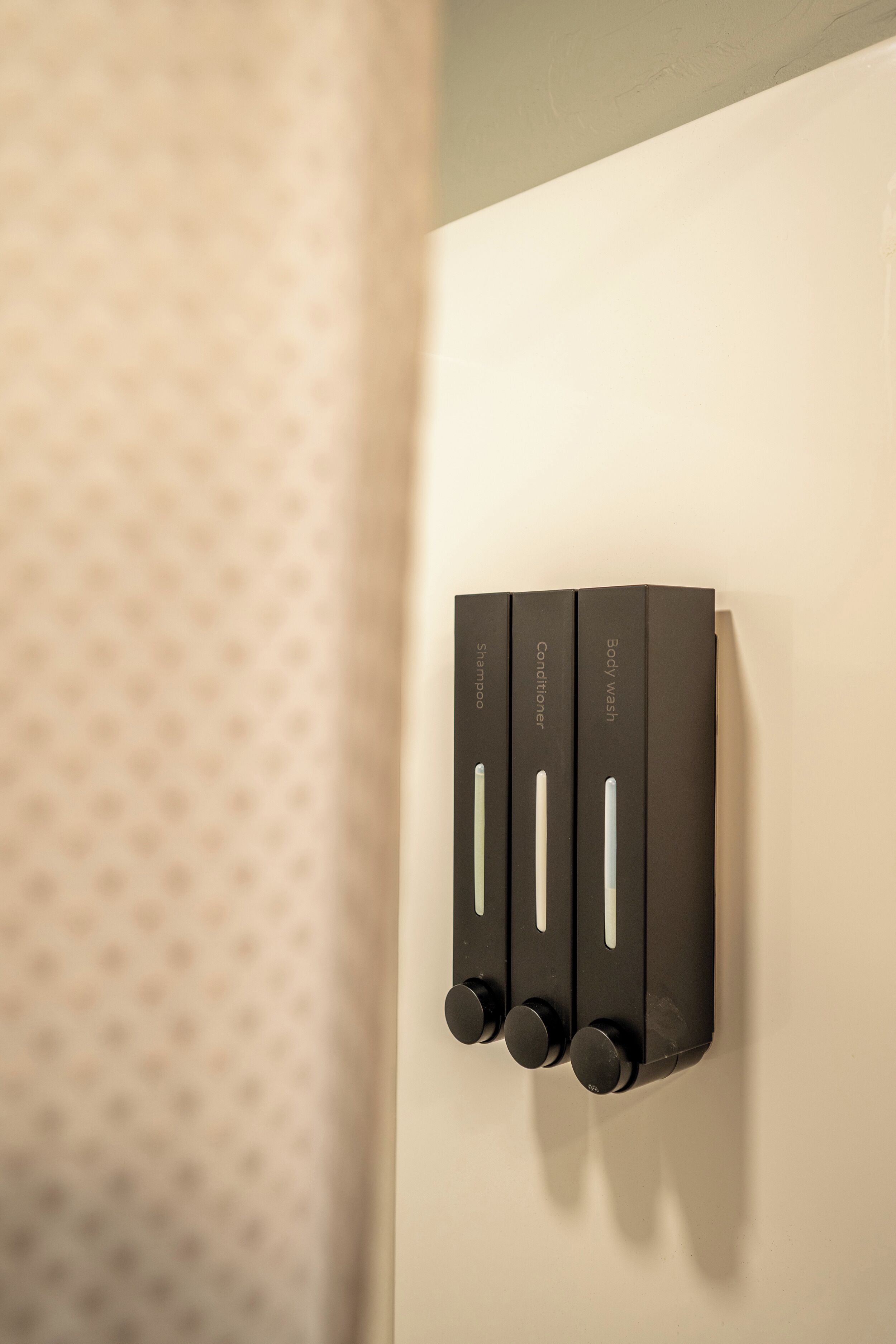 A wall-mounted dispenser with three compartments labeled 'Shampoo,' 'Conditioner,' and 'Body Wash.'
