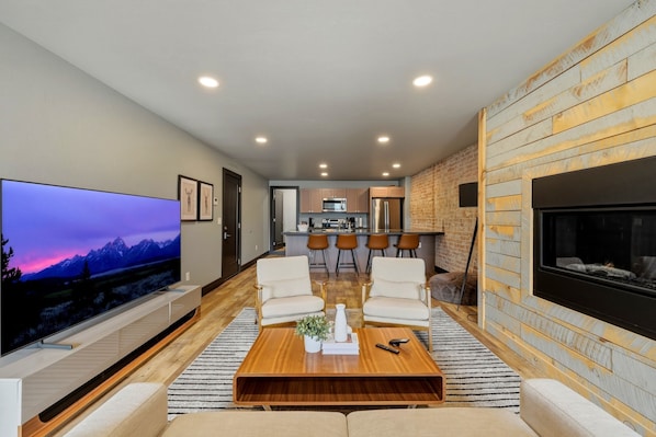 A modern living room with a flat-screen TV, a fireplace, and a kitchen in the background.