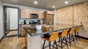 Una cocina moderna con una isla grande, taburetes de bar, pared de ladrillo y una puerta que da a otra habitación.