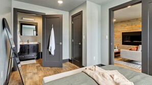 Una habitación de hotel moderna con cama, zona de estar con sofá, televisor de pantalla plana, pared decorativa de madera y baño con espejo y mueble.