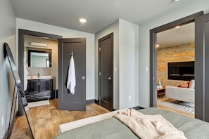 A modern hotel room with a bed, a seating area with a sofa, a flat-screen TV, a wooden accent wall, and a bathroom with a mirror and cabinet.
