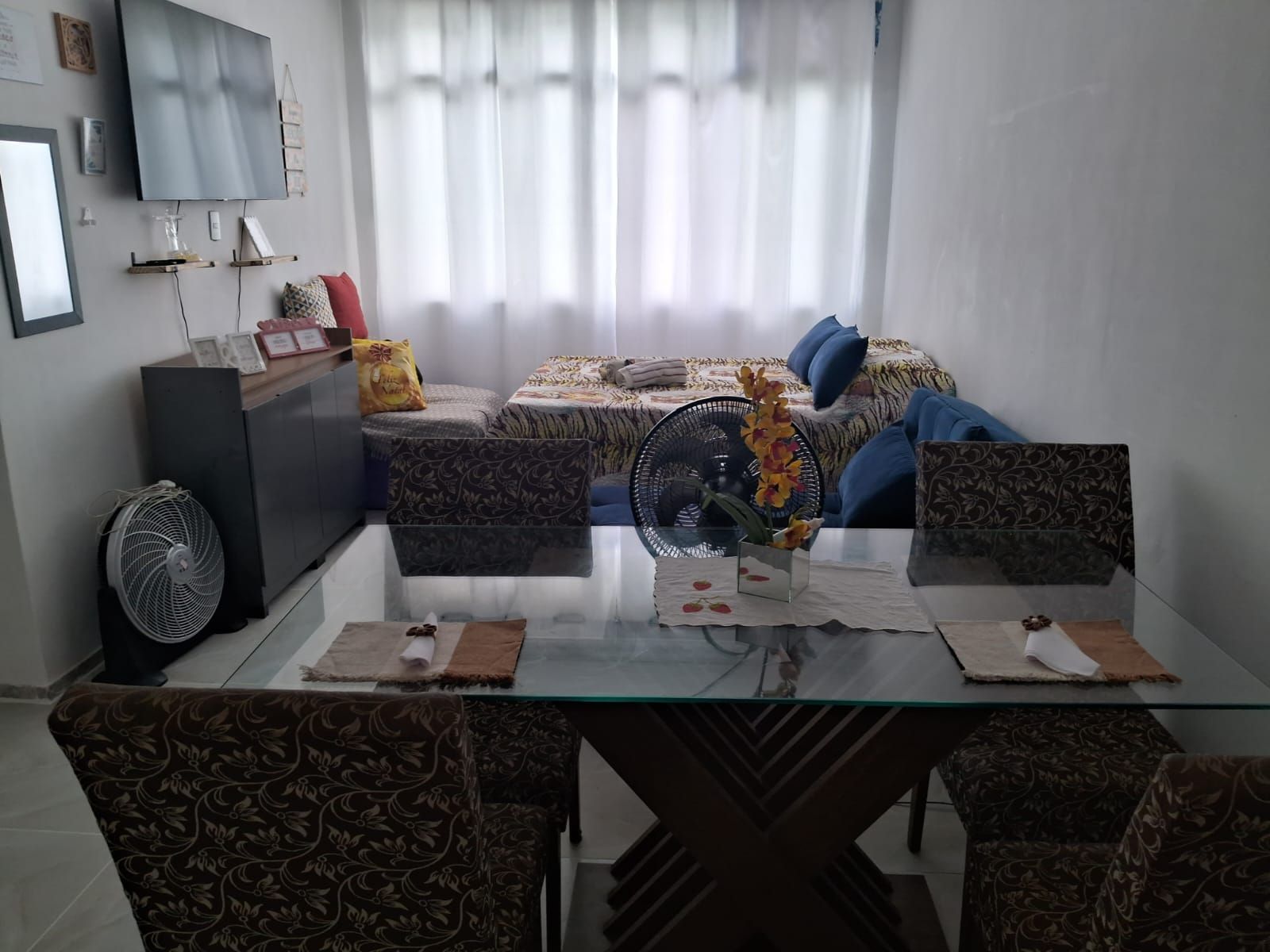 A modern living room with a glass dining table, patterned sofas, a large bed with a zebra print bedspread, and a standing fan.