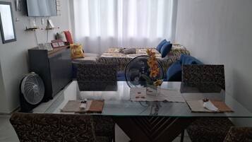 A modern living room with a glass dining table, patterned sofas, a large bed with a zebra print bedspread, and a standing fan.
