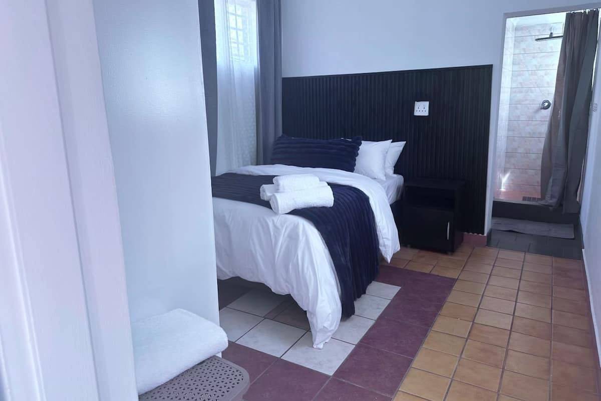 Habitación de hotel con cama, mesita de noche, un taburete pequeño, zona de ducha y suelo de baldosas.