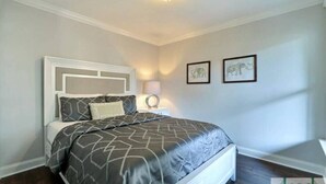 Un dormitorio con cama, mesita de noche y lámpara de pared. Se observan dos cuadros en la pared.