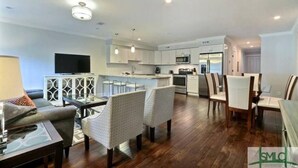 Una sala de estar moderna con un sofá, sillas y un televisor. Incluye una cocina con electrodomésticos y un comedor con sillas.