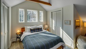 Une chambre avec un grand lit, deux fenêtres, une poutre en bois au plafond, une table de chevet avec une lampe et une porte avec des stores.