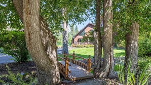Een houten bruggetje naar een huis, omringd door grote bomen.