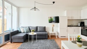 Un appartement moderne avec un canapé gris, une petite table ronde, un coin cuisine et une grande fenêtre.