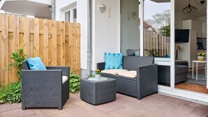 Un espace extérieur aménagé en terrasse, avec des meubles en osier, une clôture en bois et une porte-fenêtre coulissante donnant sur la cuisine.