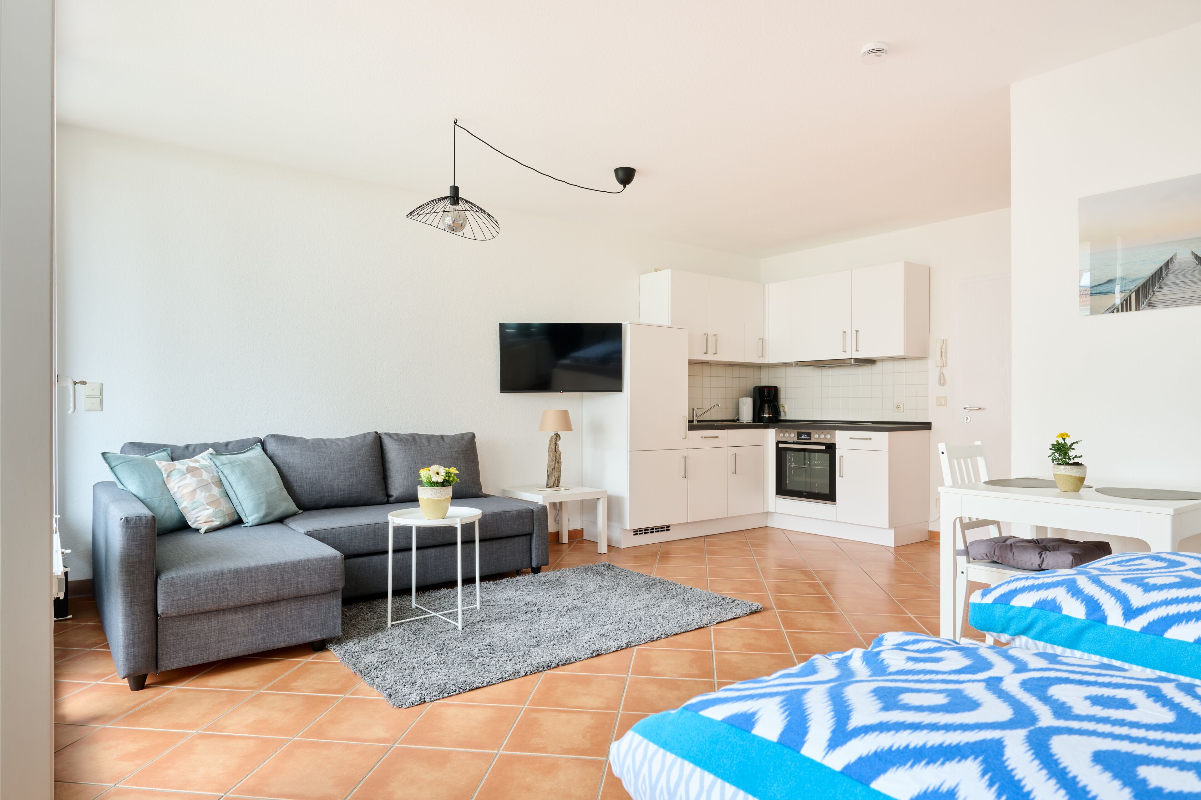 Un appartement moderne avec un canapé gris, une petite table blanche, un tapis gris, une cuisine avec des meubles blancs et un coin repas avec une table et des chaises blanches.