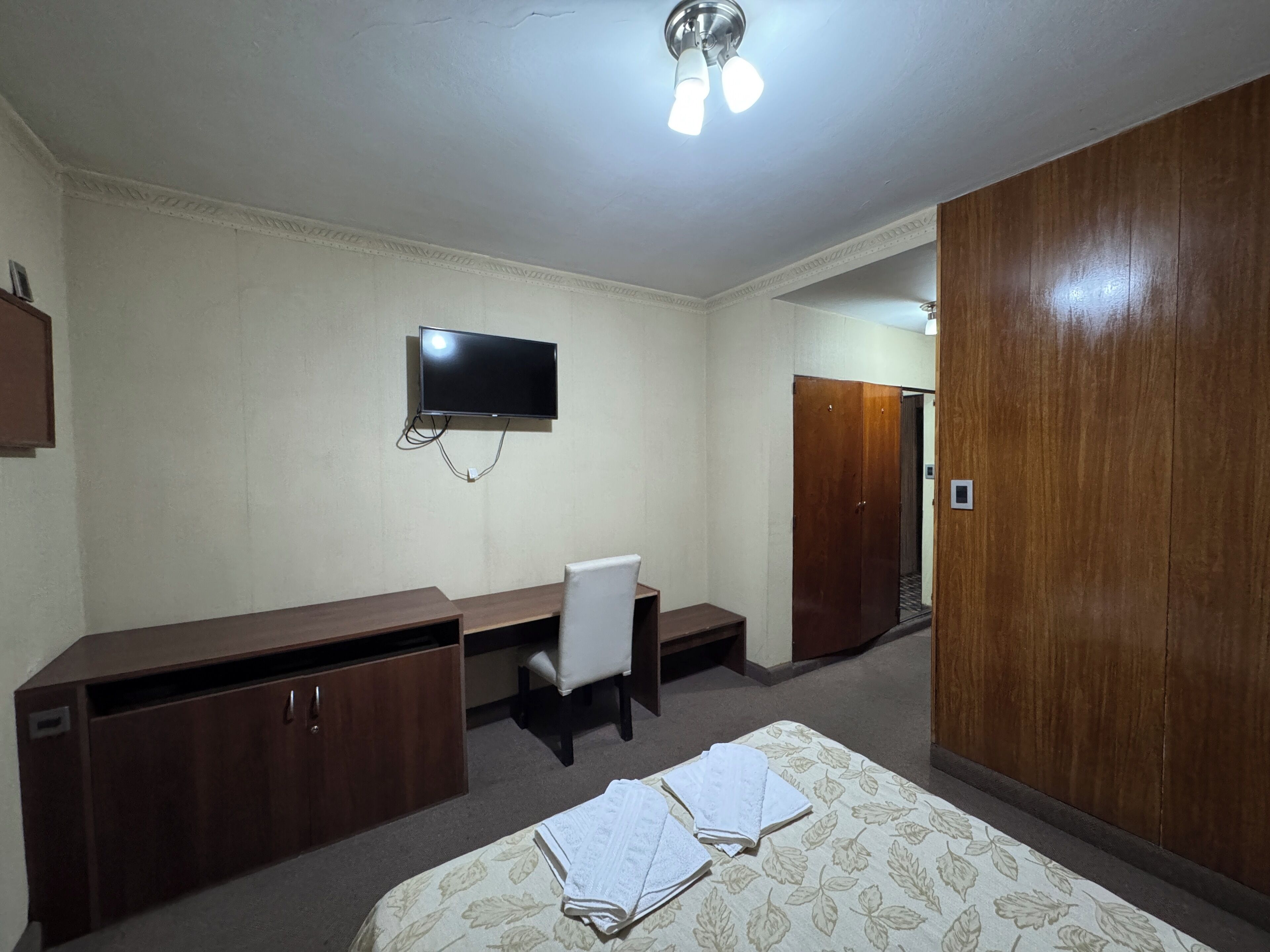 A bedroom with a bed, a desk, a chair, a wardrobe, a television, and a ceiling light.
