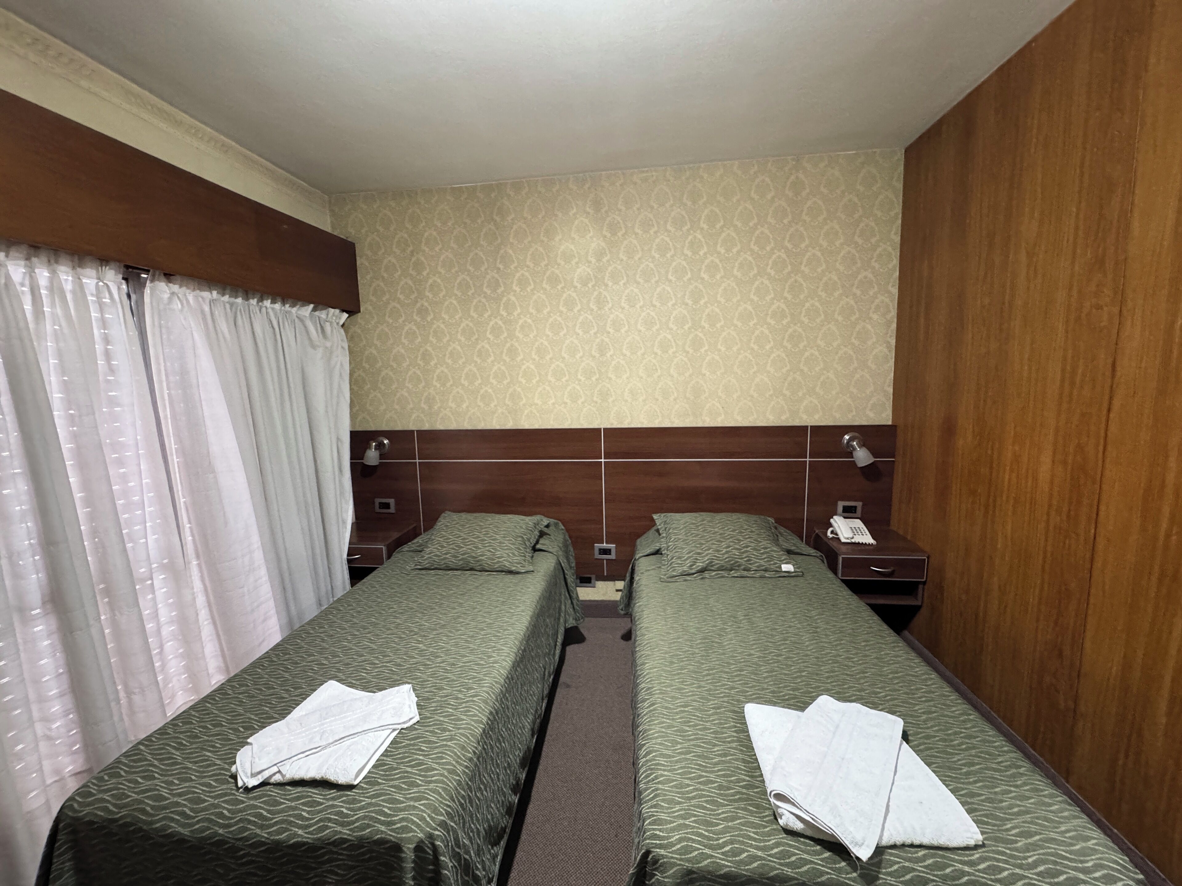 Habitación de hotel con dos camas individuales, cabecero de madera, pared con estampado y cortina.