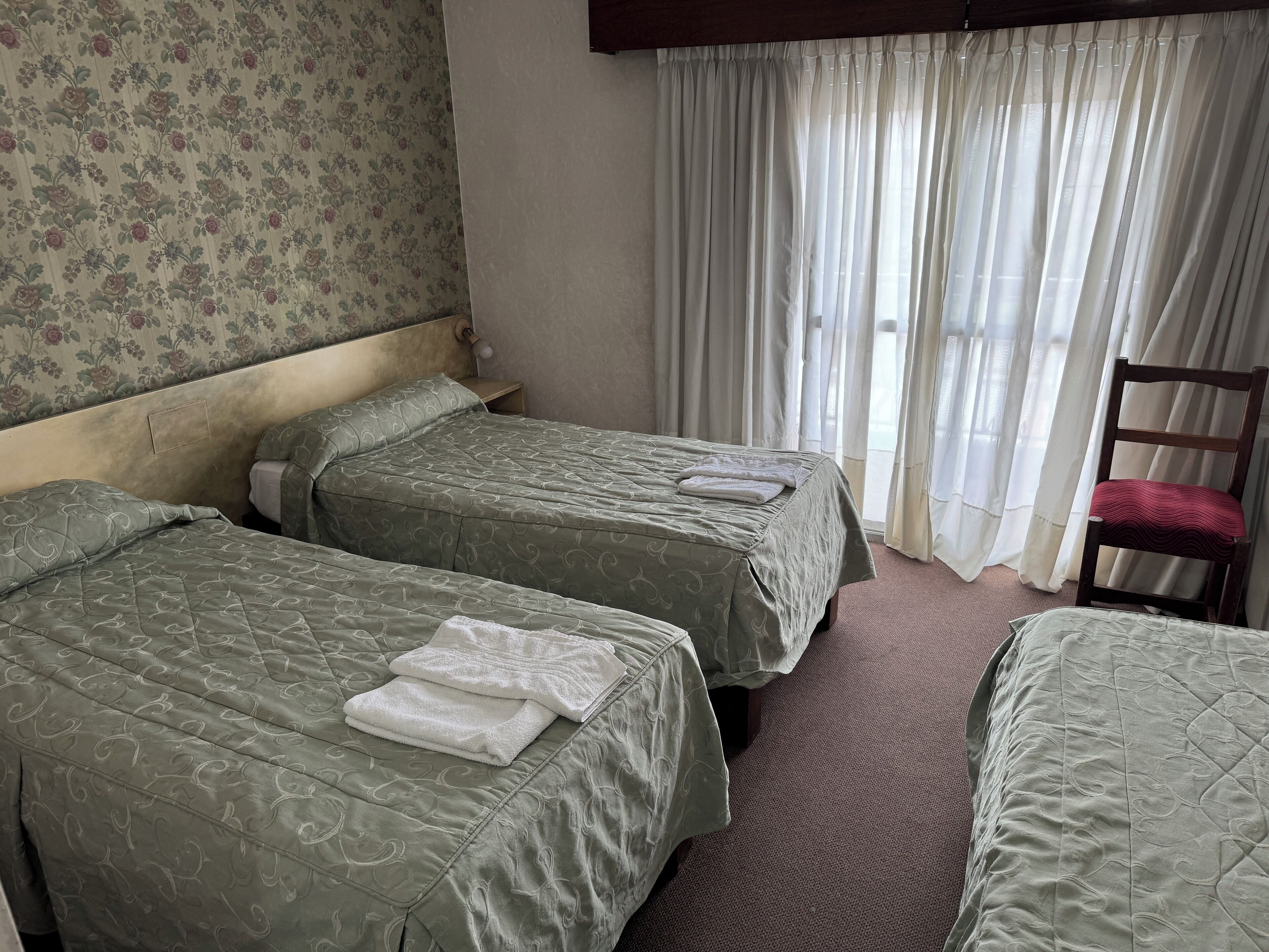 A hotel room with two beds, a wooden chair, and a window with sheer curtains.