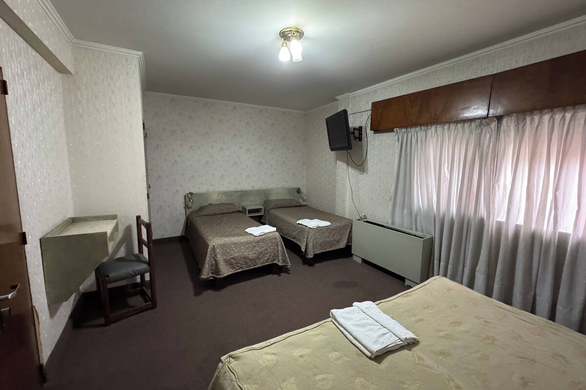 Ein Hotelzimmer mit zwei Einzelbetten, einem Schreibtisch, einem Stuhl, einem an der Wand montierten Fernseher und einem Fenster mit Vorhängen.