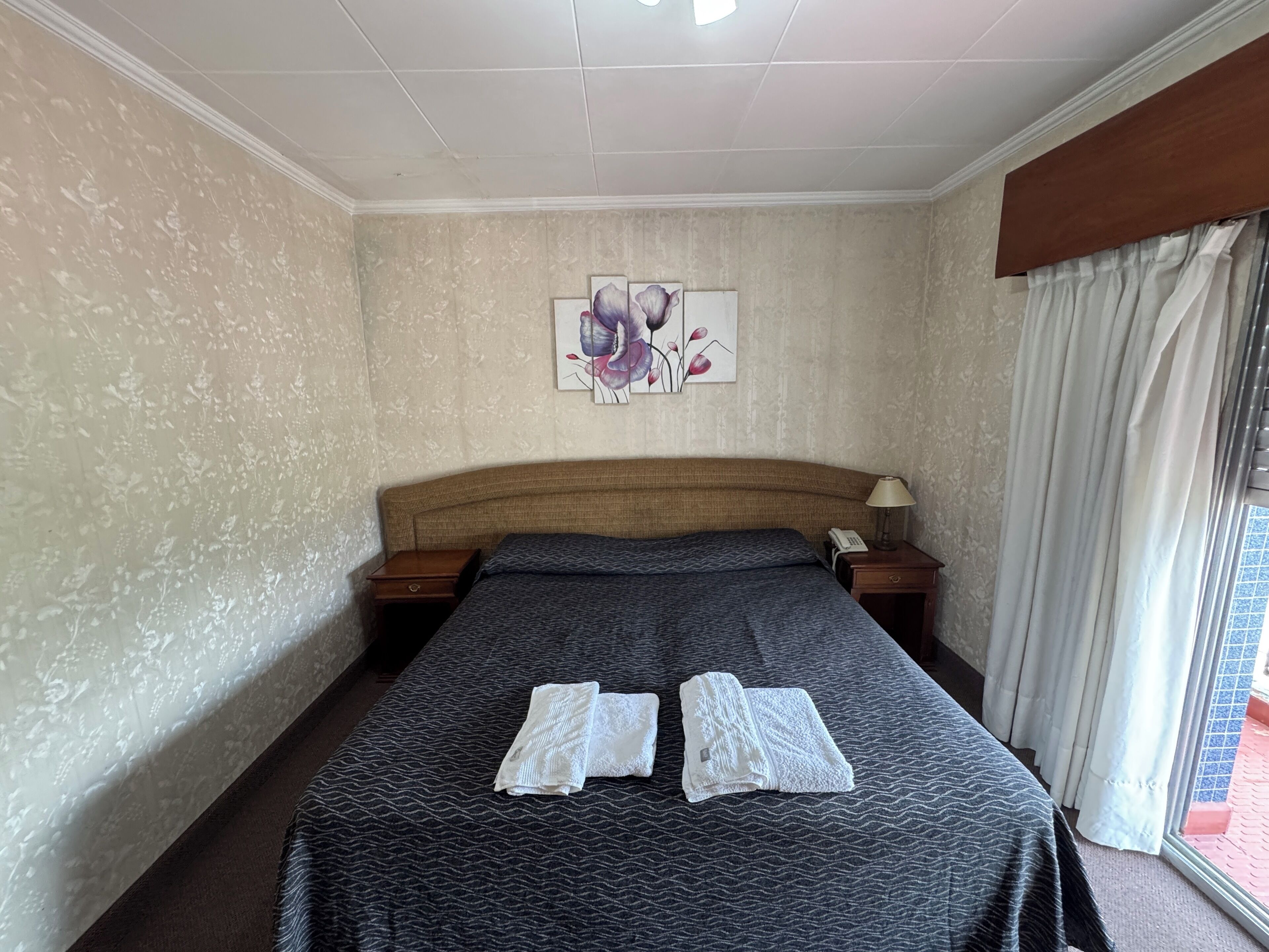 Ein Hotelzimmer mit einem großen Bett, einem Kopfteil, einem Nachttisch mit Lampe und einem Gemälde an der Wand.