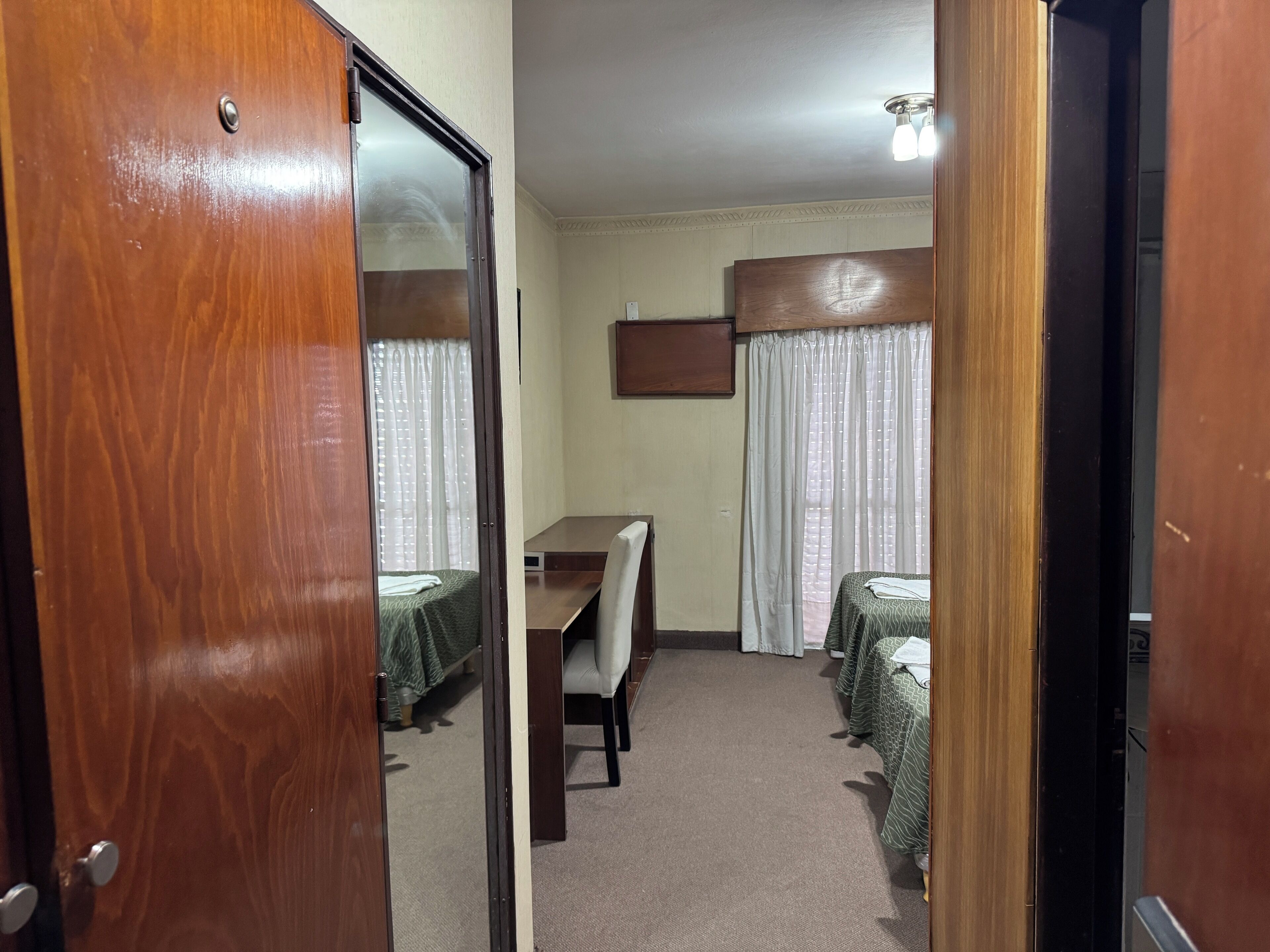 Une chambre d’hôtel avec une porte en bois, un bureau, deux lits recouverts de couvre-lits à motifs verts, une chaise et un luminaire au plafond.