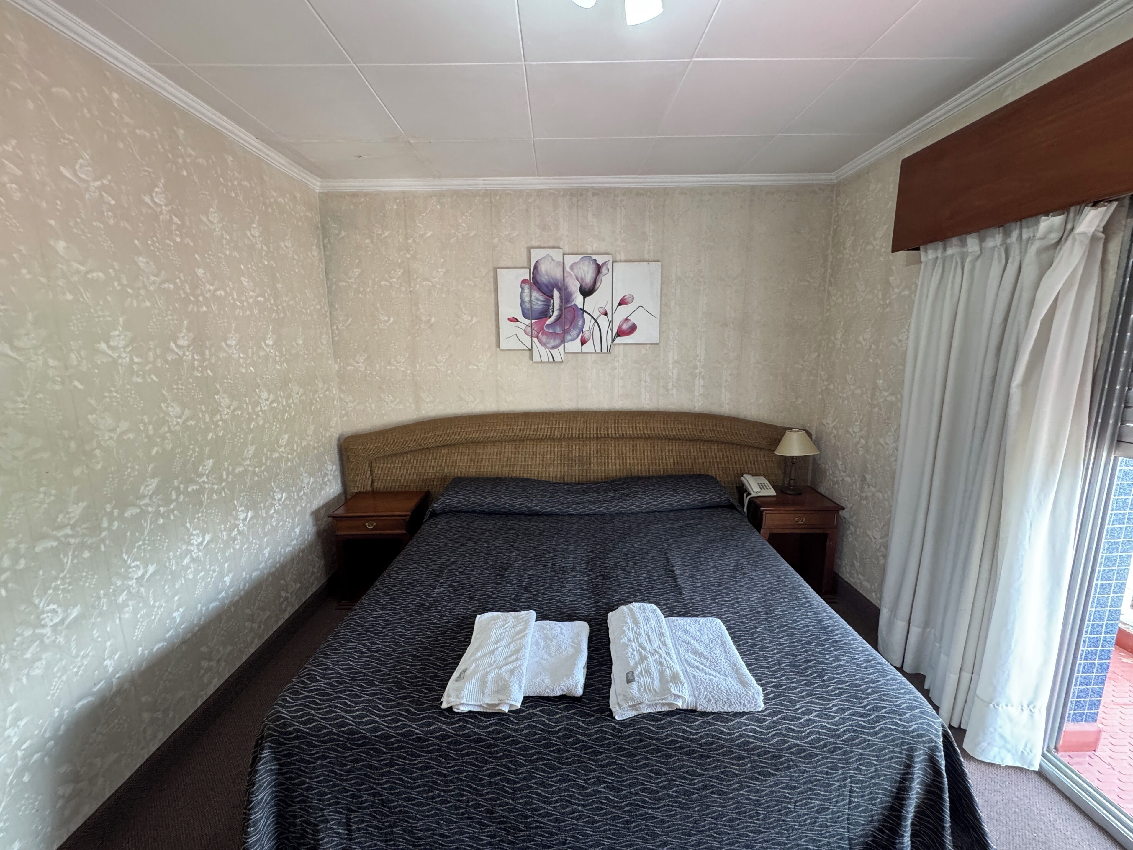 Habitació d'hotel amb un llit gran, dues taules de nit, un telèfon, un quadre a la paret i una finestra amb cortines.
