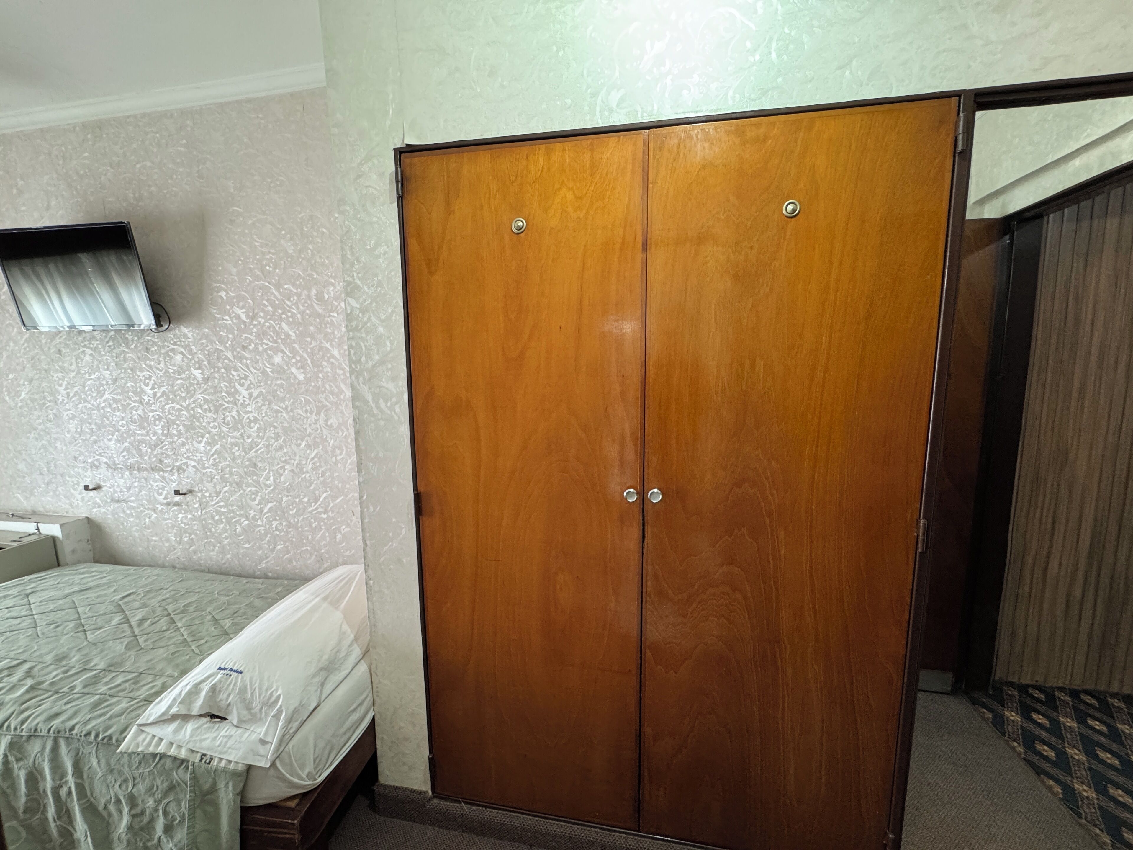 Une pièce avec une armoire en bois, un lit recouvert d’une courtepointe blanche et un téléviseur fixé au mur.