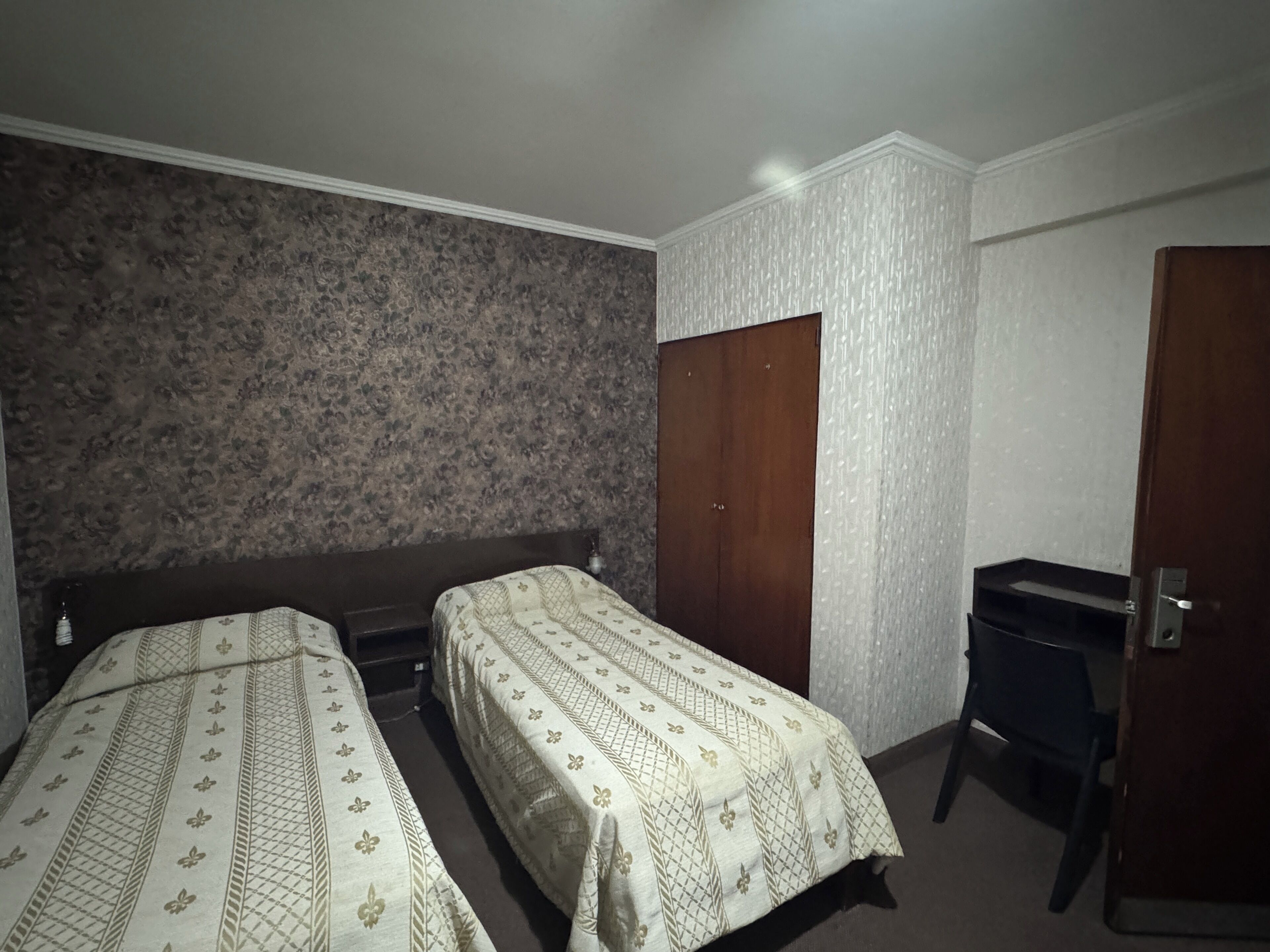 Una habitación pequeña y tenue, con dos camas individuales, un armario de madera y un escritorio.