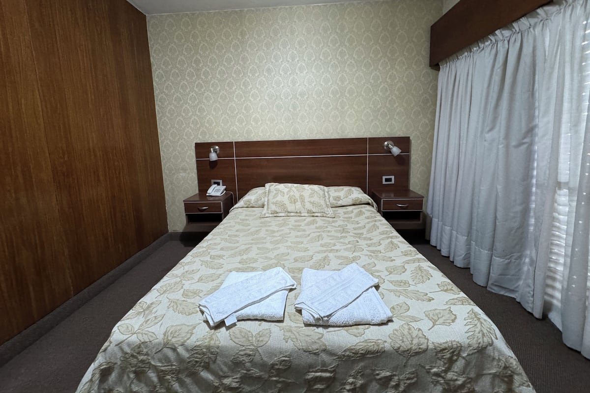 Ein Hotelzimmer mit einem großen Bett, zwei Nachttischen, einem Holz-Kopfteil, einem Telefon und einem Fenster mit Vorhängen.