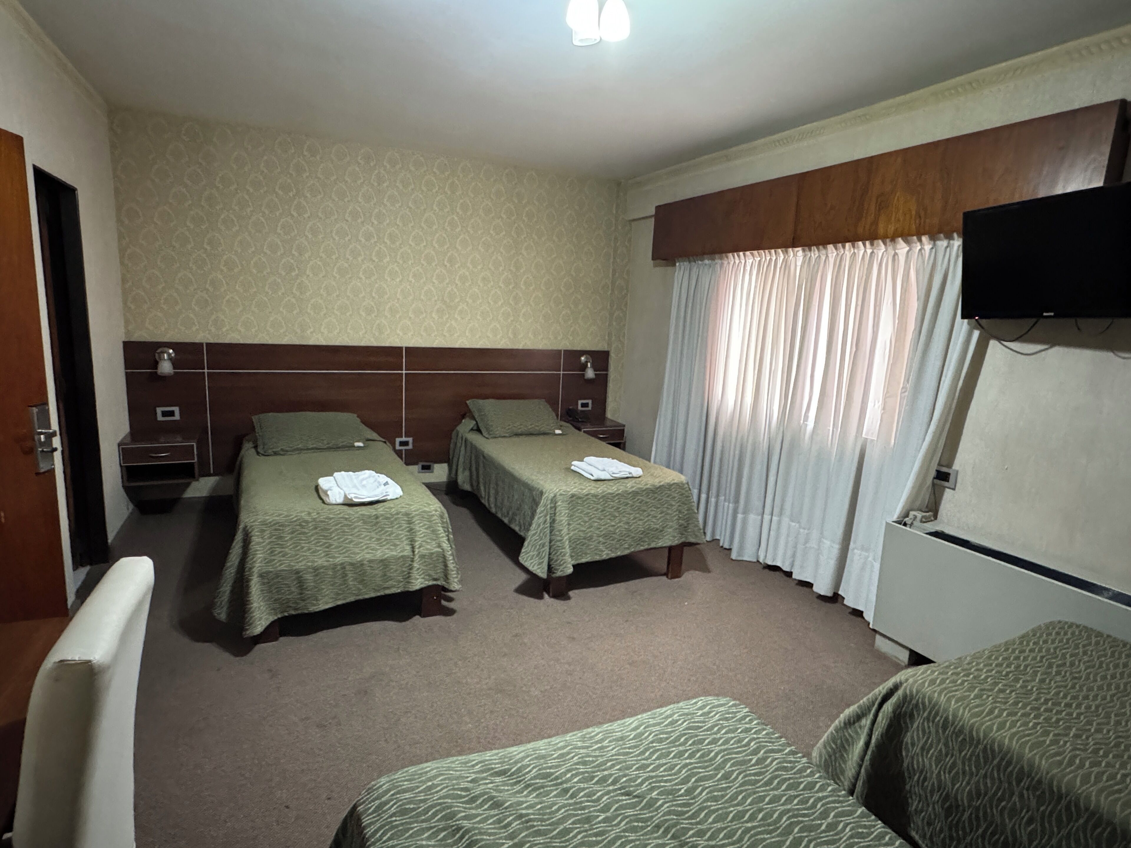 A hotel room with two single beds, a small bedside table with a lamp, a television mounted on the wall, and a window with curtains.
