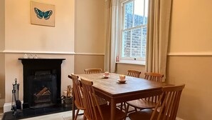 A dining room with a fireplace, wooden chairs, a table, and a window with curtains. - Snug cottage in the South Downs (Walderton)