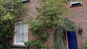A two-story brick house with a blue door, a white window, and a small garden area with various plants. - Snug cottage in the South Downs (Walderton)