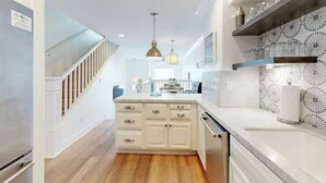 A modern kitchen with white cabinetry, a wooden floor, a built-in dishwasher, and a pendant light above the island. - Oceanfront Escape | Steps to Beach | Sleeps 7 (Carlsbad)