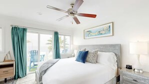 A spacious bedroom with a large bed, a ceiling fan, a view of the outdoors, and a nightstand with a lamp. - Oceanfront Escape | Steps to Beach | Sleeps 7 (Carlsbad)