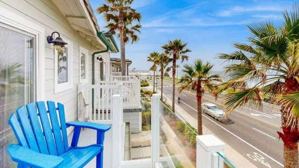 A balcony with a blue chair, a white railing, and a view of a street with palm trees. - Oceanfront Escape | Steps to Beach | Sleeps 7 (Carlsbad)