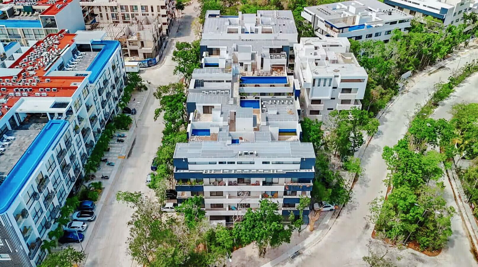 Un edificio de varios pisos con techo plano, rodeado de vegetación y al lado de una carretera.