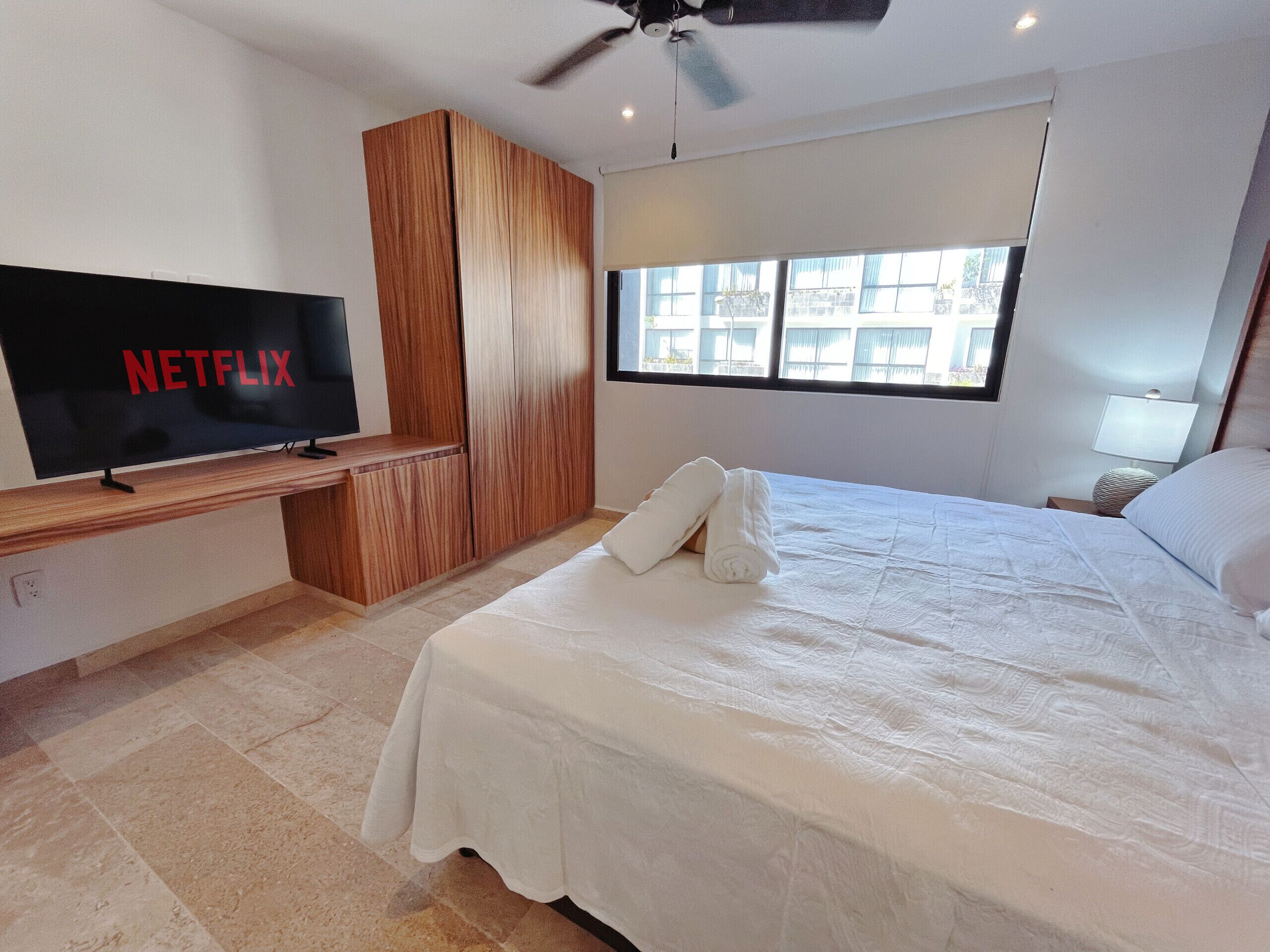 A modern hotel room with a large flat-screen TV displaying 'NETFLIX', a bed with white linens, a ceiling fan, and a window with a view of buildings.