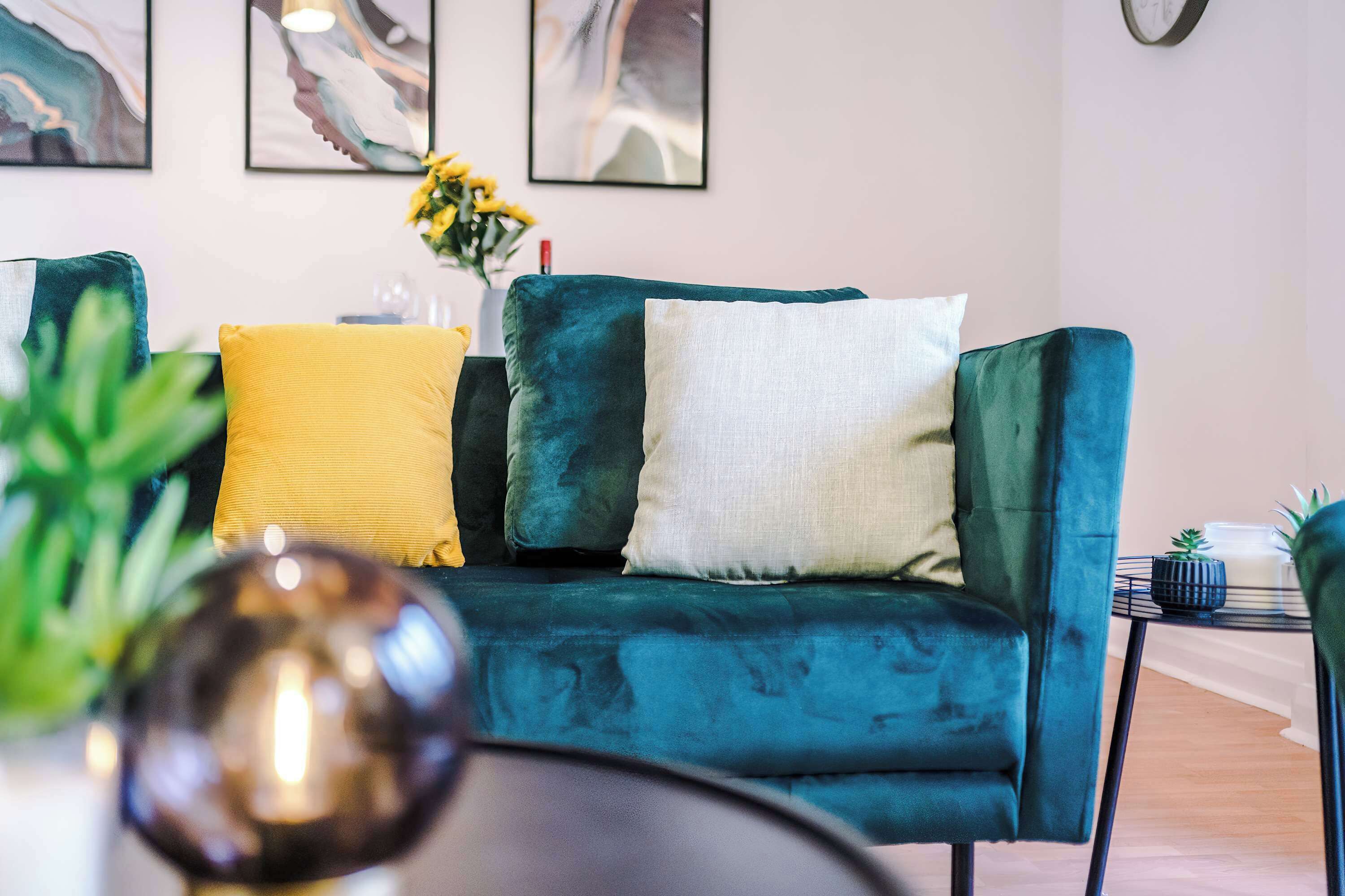Un canapé de couleur turquoise agrémenté de coussins jaunes et blancs, une petite table avec une plante, et une œuvre d’art abstrait accrochée au mur.