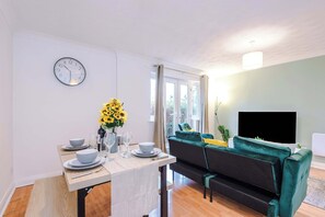 A dining area with a table set for two, a clock on the wall, and a sofa with a TV in the background. - Modern 1BR MediaCity Stay - Free Parking - M50 (Manchester)