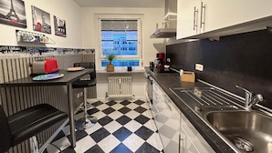A compact kitchen with a black and white checkered floor, a dining area with a table and chairs, and a large window with a view of buildings. - Cozy bright apartment centrally located with balcony and parking space (Bayreuth)