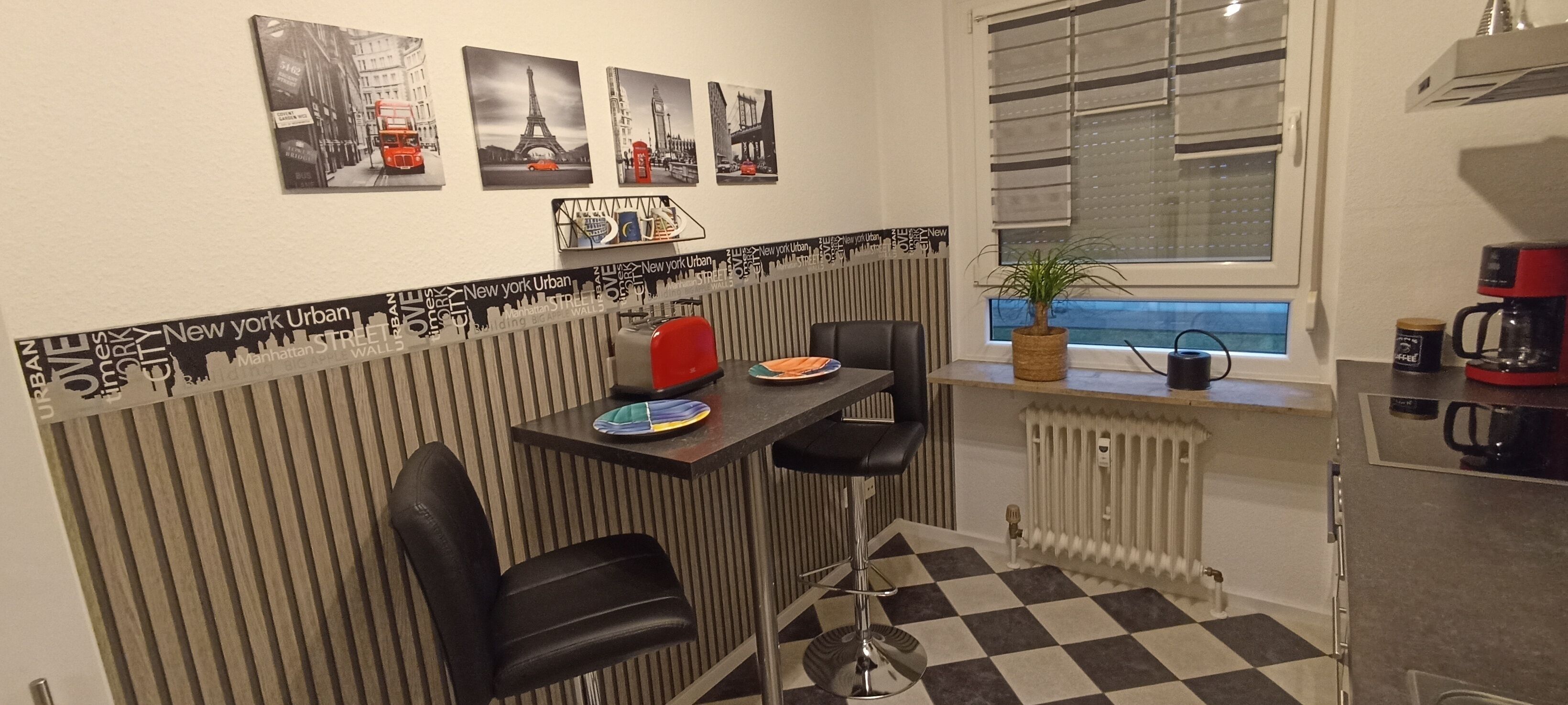 Et lille køkken med et sort-hvidt skakmønstret gulv, en radiator, en kaffemaskine og et lille bord med to stole.