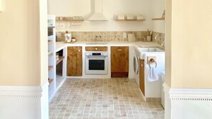 Une cuisine avec un sol carrelé, des meubles de rangement en bois, un four encastré, un lave-linge et un luminaire suspendu.