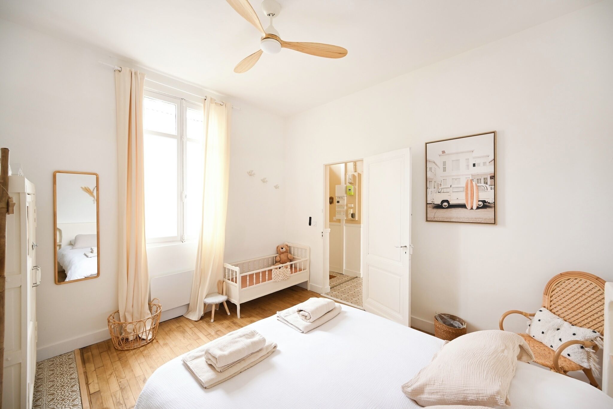 Une pièce lumineuse et blanche, avec un lit, un berceau, une chaise en osier et un ventilateur de plafond.
