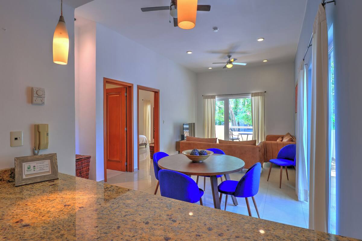 A modern kitchen with granite countertops, a dining area with blue chairs, and a living room with a sofa and a TV.