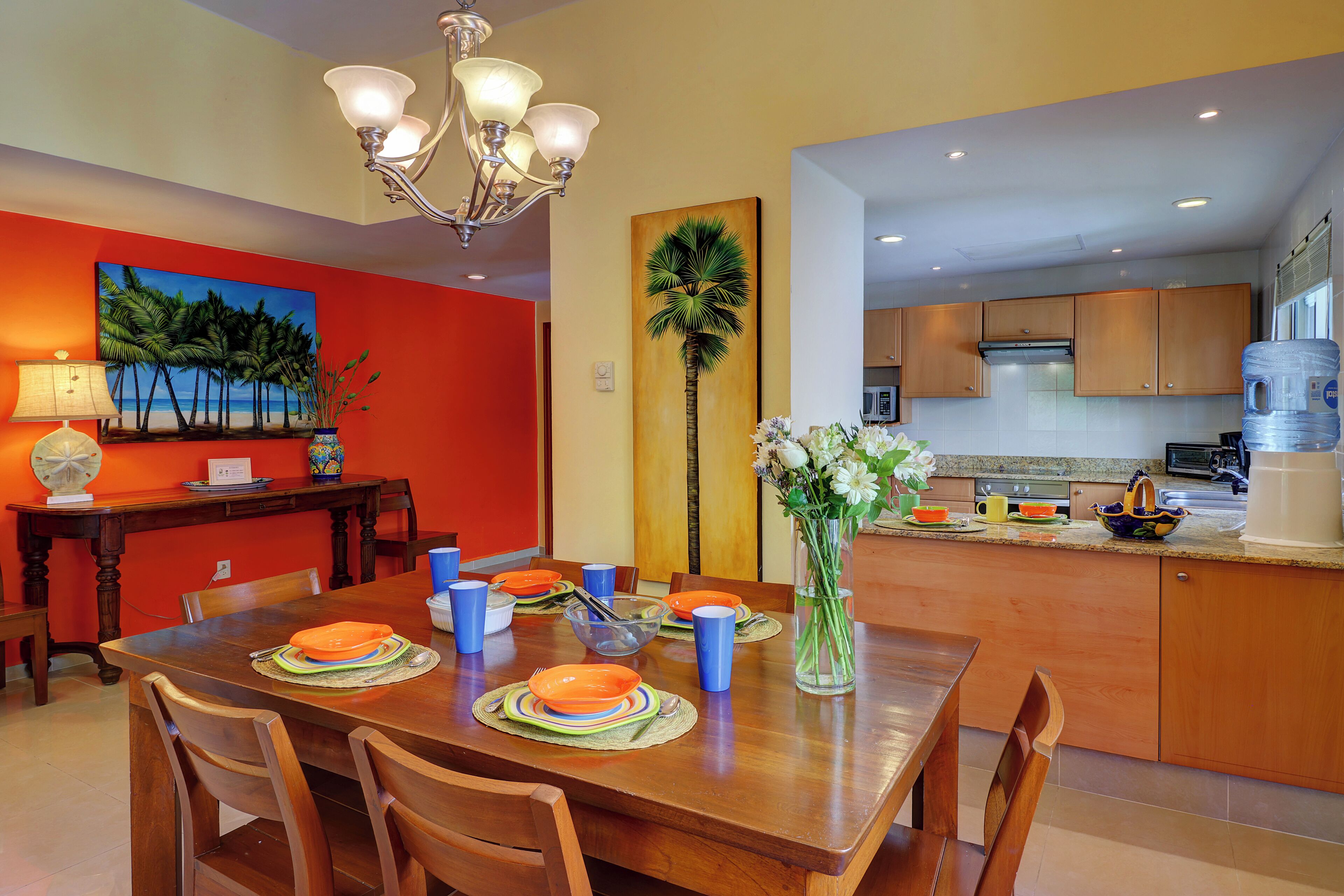 A dining area with a wooden table set for four, a vase with flowers, and a painting of palm trees on the wall.