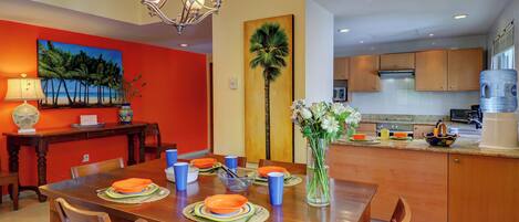 A dining area with a wooden table set for four, a vase with flowers, and a painting of palm trees on the wall.
