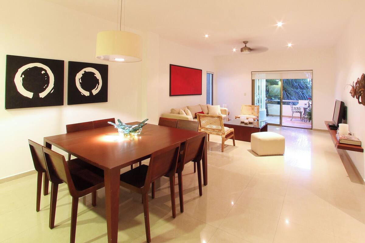 A modern living room with a dining area, a sofa set, and a large window with a view of greenery.